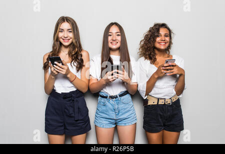 Trois belles jeunes filles portant des vêtements décontractés à la vers le haut et l'utilisation de téléphones cellulaires les plus isolées d'arrière-plan gris Banque D'Images