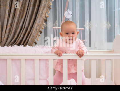 Un heureux, laughing baby girl standing dans un lit dans une chambre rose scène. Banque D'Images