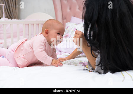 Mère avec de longs cheveux noirs joue avec son bébé enfant en vêtements rose pose à côté d'elle dans le lit Banque D'Images