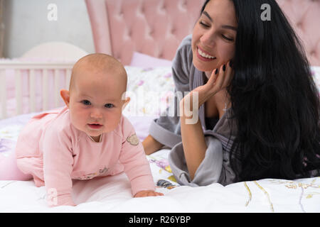 Jeune mère avec de longs cheveux noirs avec sa jeune fille au lit, regardant sa fille et sourit. L'enfant est habillé en robe rose, l'article o Banque D'Images