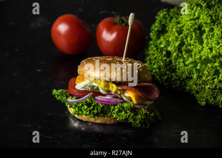 Délicieux cheesburger avec un grand nombre de légumes, moutarde, mayonnaise, de salami et d'oignon violet sur fond sombre, en bois Banque D'Images