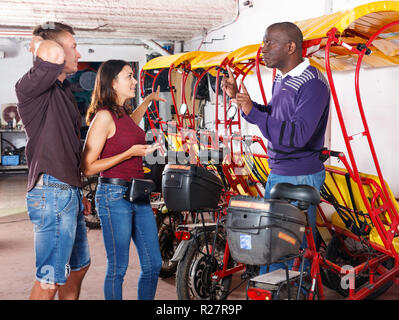 L'homme afro-américain de pilote insatisfait parle de pousse-pousse avec couple de jeunes clients souriant agréable Banque D'Images