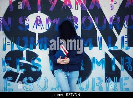 Image surréaliste comme woman student holding books et portant sur capot face invisible se pencher en arrière sur le mur de graffiti. Masque Social pour cacher son id Banque D'Images