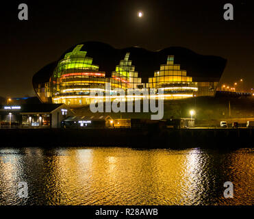 Newcastle Upon Tyne, Angleterre, Royaume-Uni, 17 novembre 2018. Vue depuis le quai le long de la rivière Tyne colorées de centre culturel Sage et salle de concert hall éclairé la nuit avec des reflets dans l'eau et la lune dans le ciel Banque D'Images