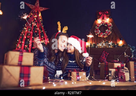 Heureuse fête de Noël, pull en bois de cerf et petite fille à santa helper hat avec baguettes en mains, coffrets cadeaux sur le premier plan. S'amuser sur salon, arbre de Noël, foyer historique Banque D'Images