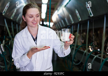 Ingénieur en production laitière femelle robe blanche debout avec verre de lait près de la ligne de traite à la ferme Banque D'Images