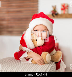 Cute young girl wearing santa hat serrant son cadeau de noël, peluche ours. Happy kid avec cadeau de Noël, en souriant et en regardant la caméra. Banque D'Images