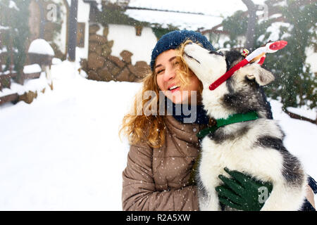 Jolie femme caucasienne hugs funny chien malamute wearing santa chers bois de noël. Female bouclés s'amusant avec chiot huskies sur nouvelle année. Le Pet est meilleur cadeau. Doggy lécher le visage de fille. Banque D'Images