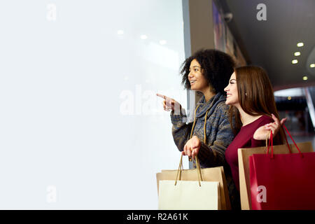 Les jeunes femmes course divers multiethnique avec des sacs éco papier près de white vitrine copyspace pour designer. Girl pointing par doigt à l'attention des passants, des vêtements et produits de mall. Banque D'Images