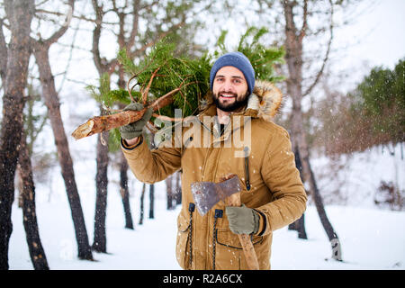 Homme barbu portant des arbres de Noël fraîchement coupés dans la forêt. Lumberjack est titulaire d'ax et sapin sur son épaule dans les bois. Comportement irresponsable envers la nature, sauver la forêt, gardez green concept. Banque D'Images