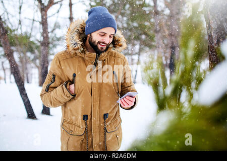 Homme barbu souriant porte des vêtements chauds pour l'hiver et l'utilisation de smartphone avec connexion internet rapide à côté de pays. Handsome man texting with cellphone et utiliser des applications en forêt. Neige dans les bois. Banque D'Images