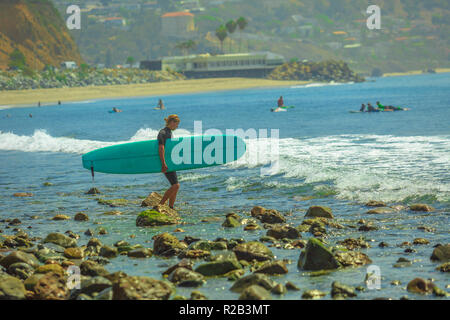 Malibu, California, United States - 7 août 2018 : surfer détient surfboard et promenades à la mer sur la plage rocheuse de Topanga Beach près de Malibu Beach en Californie à Los Angeles, célèbre pour le surf. Banque D'Images