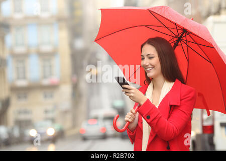 Femme heureuse en rouge en utilisant un téléphone intelligent sous la pluie en hiver dans la rue Banque D'Images
