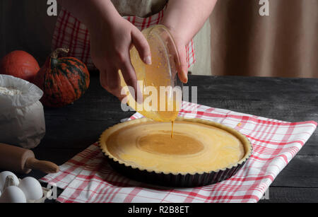 Femme mains verser la garniture pour tarte à partir d'un bol dans un bac avec de la pâte feuilletée, entouré par des ingrédients pour tarte à la citrouille. Tarte sucré classique de la cuisson. Banque D'Images