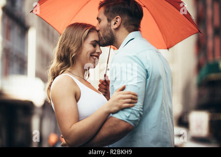Man holding sa copine et les baisers sur son front debout dans la rue. Happy couple standing in street enlacés tenant un parapluie. Banque D'Images