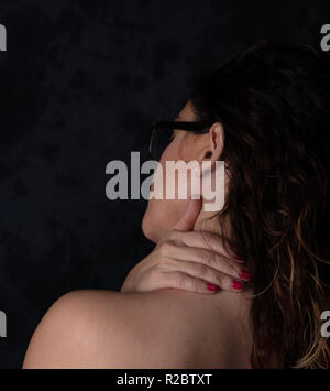 Jeune fille à la main sur son cou montrant des douleurs musculaires, photo prise à l'arrière sur le modèle noir bacground, porte des lunettes et les cheveux bruns, il n'y a fre Banque D'Images