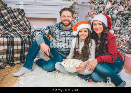 Photo de famille assis sur le plancher et souriant à la caméra. Jeune homme a la télécommande en main. Girl détient bol de maïs soufflé. Elle se penche à la mère. Banque D'Images