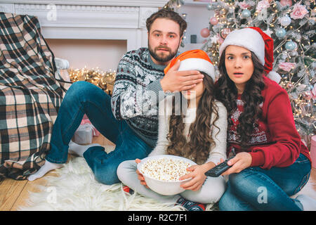 Peur parents regarder droit sur l'appareil-photo. Coveres père yeux de sa fille à la main. Ils regardent ensemble. Girl détient bol de maïs soufflé. Elle se trouve entre les parents. Banque D'Images