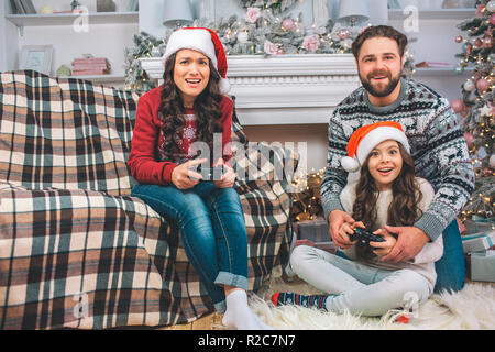 Petite fille et jeune femme joue une partie à l'aide de joysticks. Père aide sa fille. Ils s'amusent. Famille est concentré. Elles sont décorées dans les prix. Banque D'Images