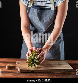 Une fille dans un tablier est titulaire d'ananas bio sur une planche en bois sur une table brune autour d'un fond sombre avec copie espace. Pour dessert ingrédient Banque D'Images