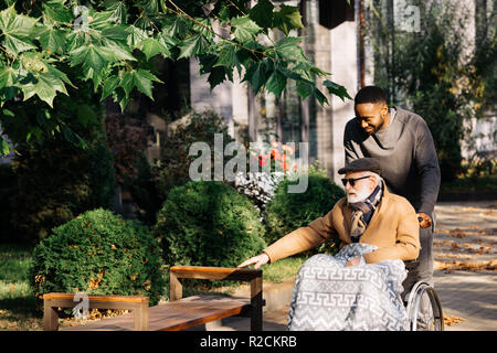 Happy senior homme handicapé en fauteuil roulant et african american cuidador de passer du temps ensemble sur la rue Banque D'Images