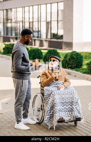 Hauts homme handicapé en fauteuil roulant avec plaid et african american man prévues tome ensemble sur rue avec les gobelets de café Banque D'Images