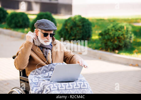 Hauts homme handicapé en fauteuil roulant à l'aide d'un ordinateur portable avec écouteurs sur street Banque D'Images