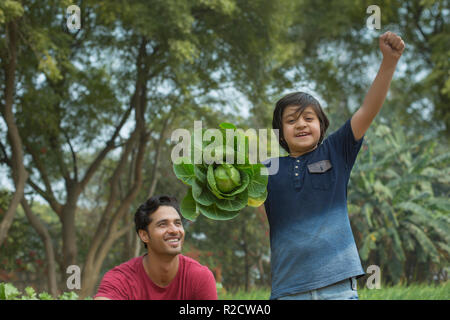 Happy Young boy standing in garden holding un gros choux dans une main et d'élever son poing avec son père assis à côté de lui. Banque D'Images