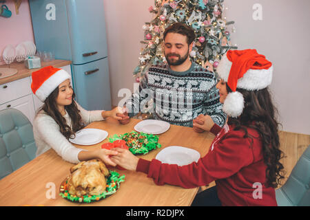 Belle et charmante les membres de la famille assis à table et de jouer. Ils tiennent les mains et garder les yeux fermés. Ils prient avant de manger la nourriture de fête. Banque D'Images