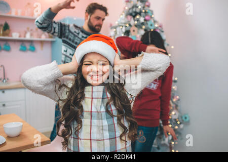 Petite fille en vêtements de fête, stands et fermer ses oreilles avec ahnds. Elle a fermé les yeux. Il y a des parents derrière elle. Le père conserve jusqu'à la main. Il va battre sa femme. Il tente de protéger elle-même. Banque D'Images