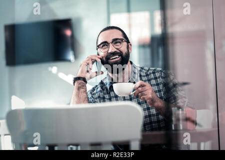 Homme barbu souriant parlant par téléphone tout en buvant un café Banque D'Images