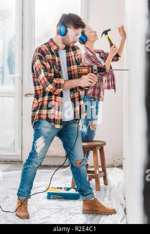 Vue latérale du jeune couple et de martelage de forage pendant la rénovation mur