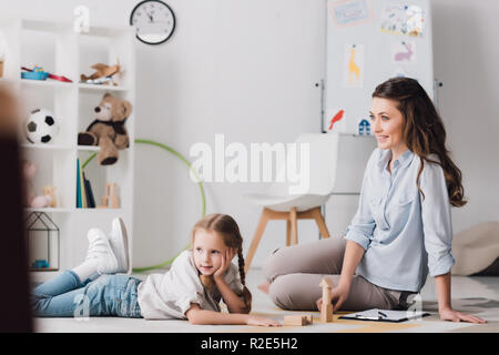 Psychologue souriant assis près de l'enfant pendant qu'elle couché sur le plancher et à l'écart Banque D'Images