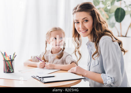 Smiling psychologue avec presse-papiers assis près de petit enfant alors qu'elle le dessin avec des crayons de couleurs et looking at camera Banque D'Images