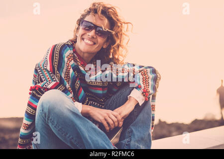 Belle Dame mignon avec les cheveux de turbulences vous sourit tandis que s'asseoir près de la côte de l'océan. poncho et vêtements hippie pour représenter la liberté concept. style de vacances dans atmosphère magique. wanderlust. Banque D'Images