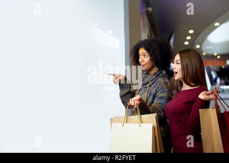 Les jeunes femmes course divers multiethnique avec des sacs éco papier près de white vitrine copyspace pour designer. Girl pointing par doigt à l'attention des passants, des vêtements et produits de mall. Banque D'Images