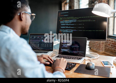 Jeune homme africain code du programme programmeur écrit assis sur le lieu de travail avec trois moniteurs dans le bureau. L'accent de l'image sur l'écran Banque D'Images