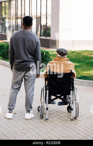 Vue arrière de la haute mobilité homme en fauteuil roulant et young african american cuidador de passer du temps ensemble sur la rue Banque D'Images