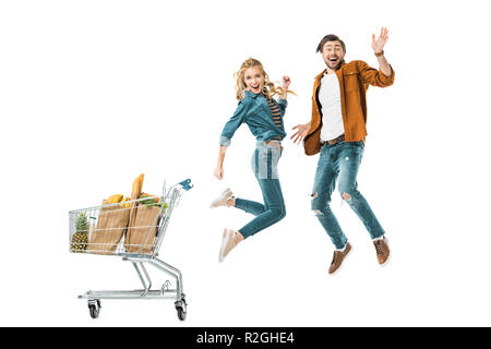 Happy young couple jumping près de panier plein de produits dans des sacs en papier isolated on white Banque D'Images