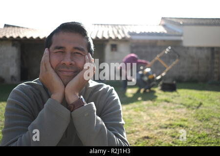 Portrait of a smiling man sitting in garden tandis qu'une femme fixe la tondeuse, France Banque D'Images