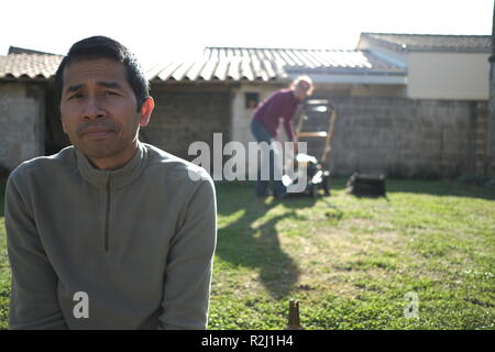 Portrait d'un homme assis dans le jardin tandis qu'une femme fixe la tondeuse, France Banque D'Images