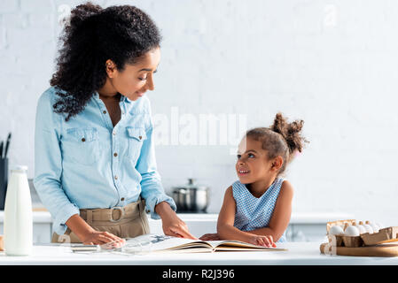 Cheerful african american mother and daughter looking at each other, livre de cuisine sur la table dans la cuisine Banque D'Images