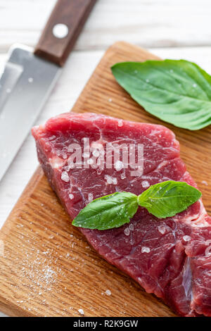 La viande marinée avec sel, poivre et basilic feuille d'assaisonnement. Matin dans la cuisine. Pavé de boeuf frais crus sur une planche à découper en bois. Fond en bois blanc, vue du dessus, copie espace, lumière du jour Banque D'Images