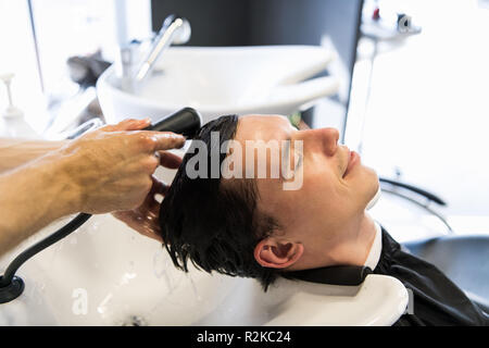 Bel homme ayant ses cheveux lavés dans de coiffure. Jeune homme couché avec ses yeux fermés en salon de beauté. Banque D'Images