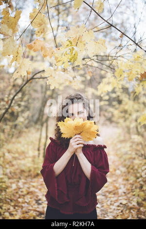 Belle jeune fille avec des cheveux noirs bouclés dans un top en soie marron dans un parc en automne, souriant à la caméra et la tenue de feuilles d'érable en face de son visage Banque D'Images