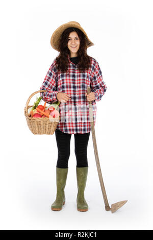 Jeune agriculteur avec un bras tenant un panier avec des fruits et légumes frais, avec l'autre une houe, elle porte un chapeau de paille, chemise à carreaux et de caoutchouc vert Banque D'Images