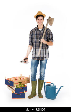 Jeune agriculteur avec cisailles à la main et pioche sur son épaule, à ses pieds avec des caisses de fruits et de légumes, il porte un chapeau de paille, une chemise à carreaux Banque D'Images