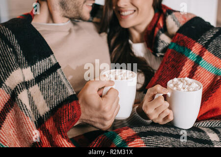 Cropped shot of smiling young couple avec tasses de cacao avec de la guimauve relaxing at home plaid sous Banque D'Images