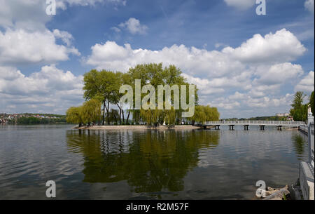 Un pont mène à une île au milieu d'un lac sur lequel de grands arbres verts croître contre un ciel bleu avec des nuages Banque D'Images