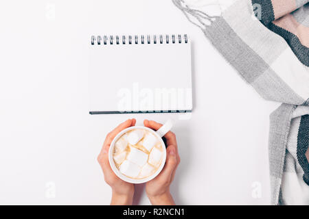 Femme's hands holding tasse de café avec des guimauves sur fond blanc à côté d'ouvrir le bloc-notes en blanc et foulard à damiers, vue du dessus. Télévision lay cdm confortable Banque D'Images